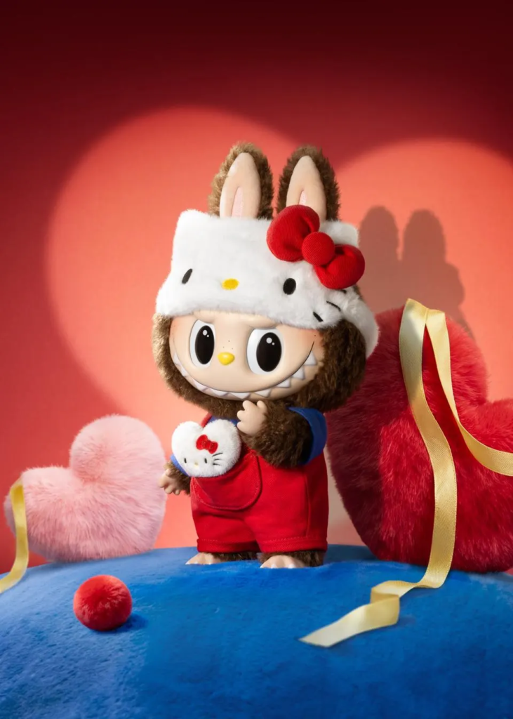 A large Labubu plush wearing a Hello Kitty hood and red overalls, styled for a Valentine's/red-themed photoshoot with heart props.